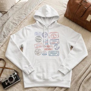 May include: White hooded sweatshirt with a kangaroo pocket, featuring travel-themed stamp designs in red, blue, and gray. Stamps include locations like London, Paris, and Prague. A vintage camera and a brown leather travel bag are also visible.
