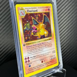 May include: A Charizard Pokémon card in a protective case. The card features a yellow and orange dragon with blue wings. The card text includes "Charizard", "120 HP", and "Fire Spin". The card is 5' 7" in length and weighs 200 lbs.