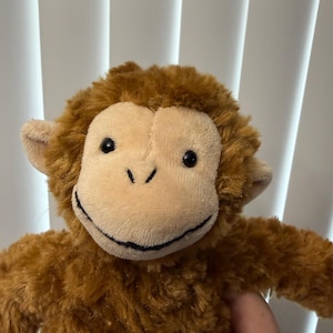 May include: A plush monkey toy featuring shaggy brown fur, a smooth beige face, small rounded ears, and black plastic eyes. The monkey has a stitched black mouth line that creates a subtle smile, with a soft, textured body and limbs.