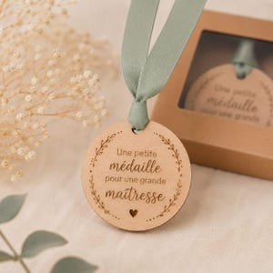 Peut inclure: Une médaille ronde en bois avec le texte "Une petite médaille pour une grande maîtresse" et un cœur. La médaille est attachée à un ruban vert sauge. Une médaille similaire est visible dans une boîte en carton.