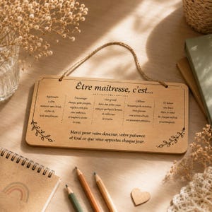 May include: A wooden plaque with the text "&Ecirc;tre ma&icirc;tresse, c'est..." in black. The plaque has a decorative floral design and a jute rope for hanging. The text is in French and is a message for teachers.
