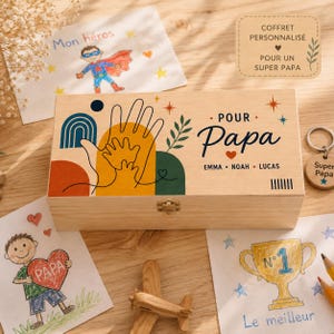 Può includere: Una scatola rettangolare in legno con una stampa che raffigura l'impronta di una mano adulta sovrapposta a quella di un bambino. Il testo recita "POUR Papa" con i nomi "EMMA • NOAH • LUCAS" sottostanti. Include una chiusura in metallo.