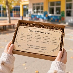 May include: A wooden plaque with a light brown finish, presented in a cardboard box. The plaque has a rope for hanging and features French text and decorative floral accents. The text is a message of appreciation.