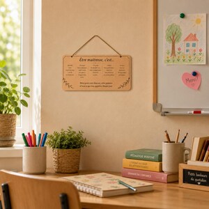 May include: A wooden sign with the text "&Ecirc;tre maitresse, c'est..." hangs on a wall. The sign is surrounded by a desk with books, pens, and a small plant. A drawing of a house and tree is on a whiteboard.