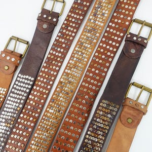 Op de afbeelding: Een collectie van zes leren riemen in verschillende bruintinten, elk voorzien van een rechthoekige messingkleurige gesp en rijen metalen studs. De riemen zijn diagonaal gerangschikt en tonen verschillende dichtheden en patronen van studs.
