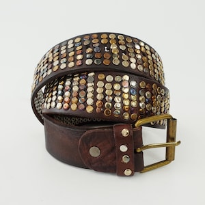 May include: A dark brown leather belt featuring multiple rows of circular metal studs in various metallic finishes including brass, silver, and copper. The belt includes a rectangular brass-toned metal buckle and a matching metal prong closure.