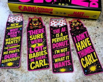 May include: Four rectangular bookmarks with black backgrounds, neon pink and yellow text, and illustrations of a cat wearing a crown and white boxers with red hearts. The text includes phrases like New Achievement, There Sure Were A Lot Of Babies In There, The AI Is A Pervert, Donut, and I Don't Have Thumbs, Carl! The bookmarks are positioned below a book titled Carl's Doomsday Scenario.