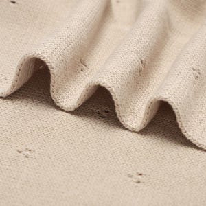 Pode incluir: Um tecido de malha bege com um padr&atilde;o pointelle subtil e pequenos orif&iacute;cios espa&ccedil;ados uniformemente. O t&ecirc;xtil apresenta uma superf&iacute;cie macia e texturizada com dobras e drapeados suaves, exibindo a trama consistente e a cor castanho-clara.
