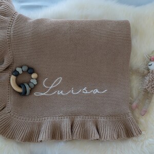 May include: A tan knitted baby blanket with a ruffled edge, embroidered with the name "Luisa" in white script. A wooden and silicone teether and a plush deer toy are also present.