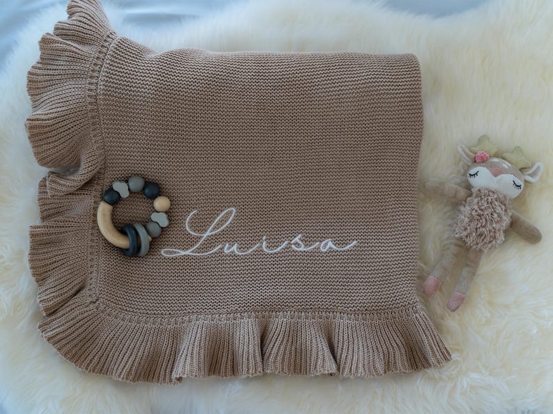 May include: A tan knitted baby blanket with a ruffled edge, embroidered with the name "Luisa" in white script. A wooden and silicone teether and a plush deer toy are also present.