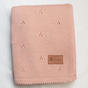 May include: A folded dusty pink knit blanket featuring a repeating pattern of small eyelet holes. A rectangular brown faux leather patch is stitched onto the lower right corner, displaying the embossed text Lenayou Collection with a floral icon.