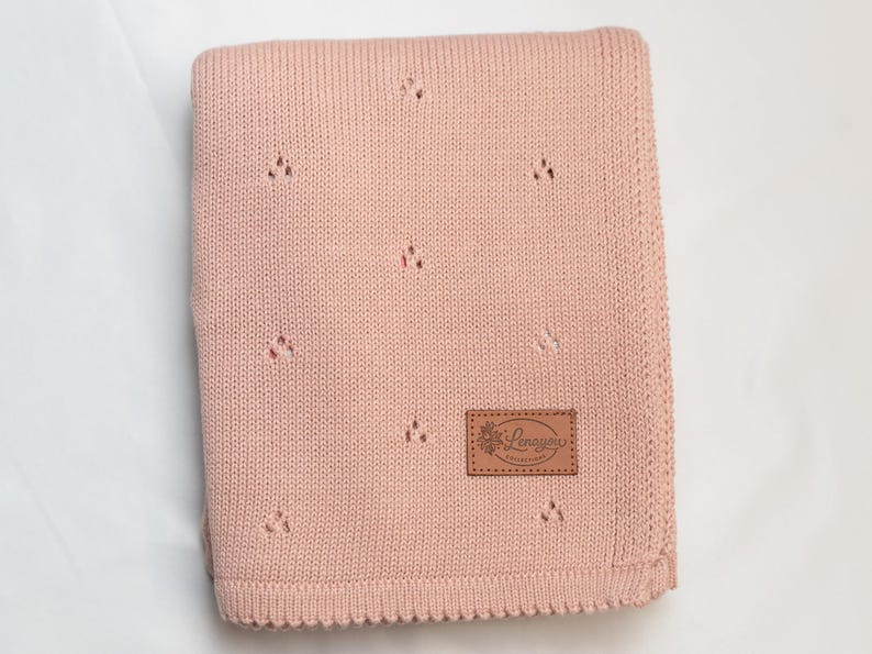 May include: A folded dusty pink knit blanket featuring a repeating pattern of small eyelet holes. A rectangular brown faux leather patch is stitched onto the lower right corner, displaying the embossed text Lenayou Collection with a floral icon.