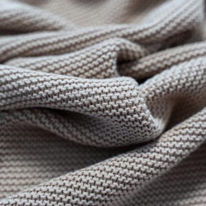 May include: Close-up of a beige knitted fabric with a textured, woven pattern. The fabric is folded and draped, showing the detailed stitch work and the soft, neutral color. The image highlights the texture and drape of the material.