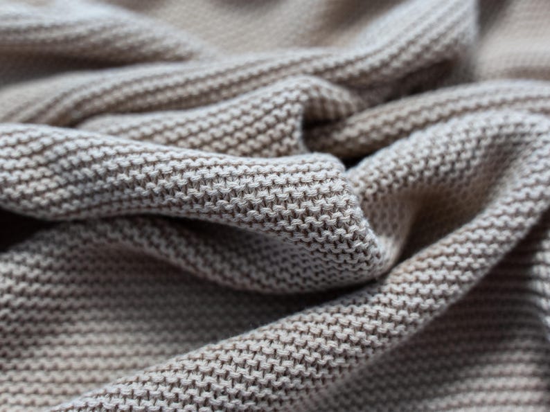 May include: Close-up of a beige knitted fabric with a textured, woven pattern. The fabric is folded and draped, showing the detailed stitch work and the soft, neutral color. The image highlights the texture and drape of the material.