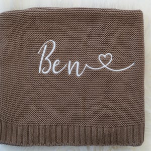 May include: A brown knitted blanket with the name "Ben" embroidered in white script, followed by a heart symbol. The blanket has a ribbed border and a soft, textured appearance.