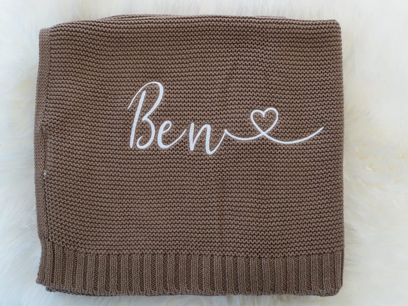 May include: A brown knitted blanket with the name "Ben" embroidered in white script, followed by a heart symbol. The blanket has a ribbed border and a soft, textured appearance.