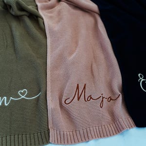 May include: Three knitted blankets in olive green, dusty rose, and black. Each blanket has a name embroidered on it in a contrasting color. The olive green blanket has the name "Ben" with a heart. The dusty rose blanket has the name "Maja". The black blanket has the name "Eliva".