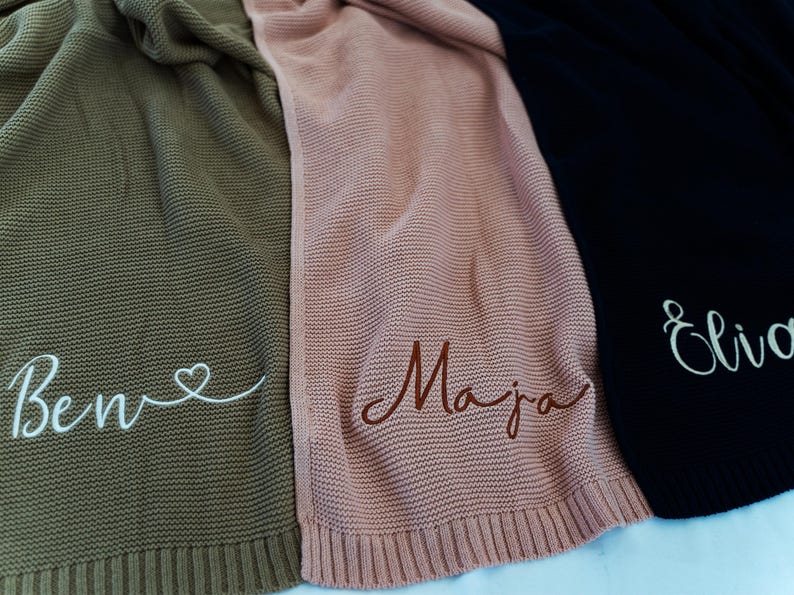 May include: Three knitted blankets in olive green, dusty rose, and black. Each blanket has a name embroidered on it in a contrasting color. The olive green blanket has the name "Ben" with a heart. The dusty rose blanket has the name "Maja". The black blanket has the name "Eliva".