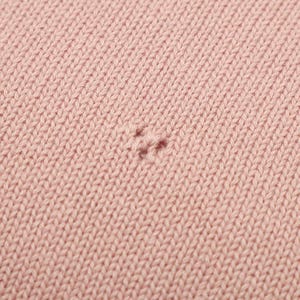 May include: A close-up view of a light pink knitted fabric featuring a consistent stockinette stitch pattern. In the center of the frame, there is a small, visible imperfection where the yarn has a slight snag or a minor pull in the knit structure.