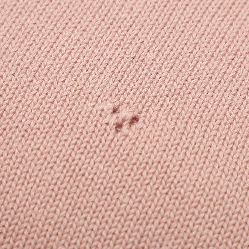 May include: A close-up view of a light pink knitted fabric featuring a consistent stockinette stitch pattern. In the center of the frame, there is a small, visible imperfection where the yarn has a slight snag or a minor pull in the knit structure.
