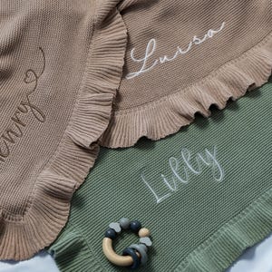 May include: Three personalized baby blankets in tan, green, and white, each with a name embroidered in a cursive font. The tan blanket says "Henry", the white blanket says "Luisa", and the green blanket says "Lilly". A wooden and silicone teether is also visible.