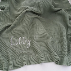 May include: Sage green knitted baby blanket with a ruffled edge. The blanket has the name "Lilly" embroidered in white script. The blanket is folded and draped, showcasing its texture and design.
