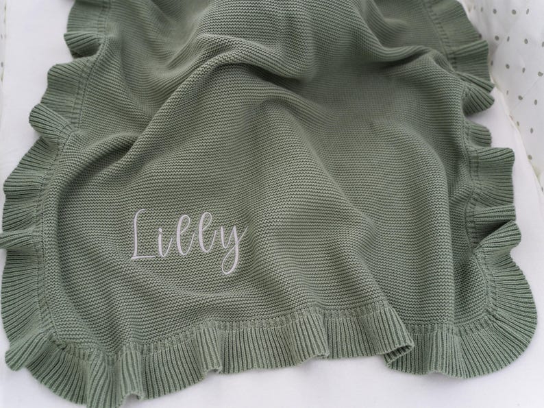 May include: Sage green knitted baby blanket with a ruffled edge. The blanket has the name "Lilly" embroidered in white script. The blanket is folded and draped, showcasing its texture and design.