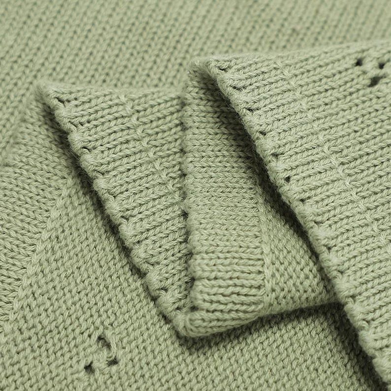 May include: A sage green knitted fabric featuring a dense, uniform stockinette stitch pattern. The material is folded to reveal multiple layers, showcasing the consistent texture, the soft matte finish of the yarn, and the clean, ribbed edges.