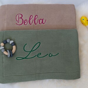 May include: Two knitted baby blankets, one tan and one green, with embroidered names "Bella" and "Leo". A wooden and silicone teething ring and a yellow maraca are next to the blankets. The blankets are on a white, fluffy surface.