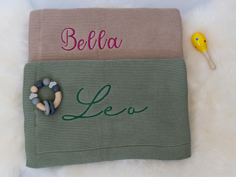 May include: Two knitted baby blankets, one tan and one green, with embroidered names "Bella" and "Leo". A wooden and silicone teething ring and a yellow maraca are next to the blankets. The blankets are on a white, fluffy surface.