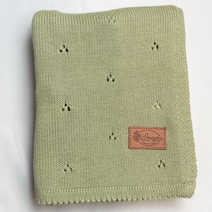 May include: A sage green knitted blanket featuring a repeating pattern of small, three-hole eyelet clusters. A rectangular brown leather-like patch is stitched onto the lower right corner, displaying the embossed text Lenayou Collections.