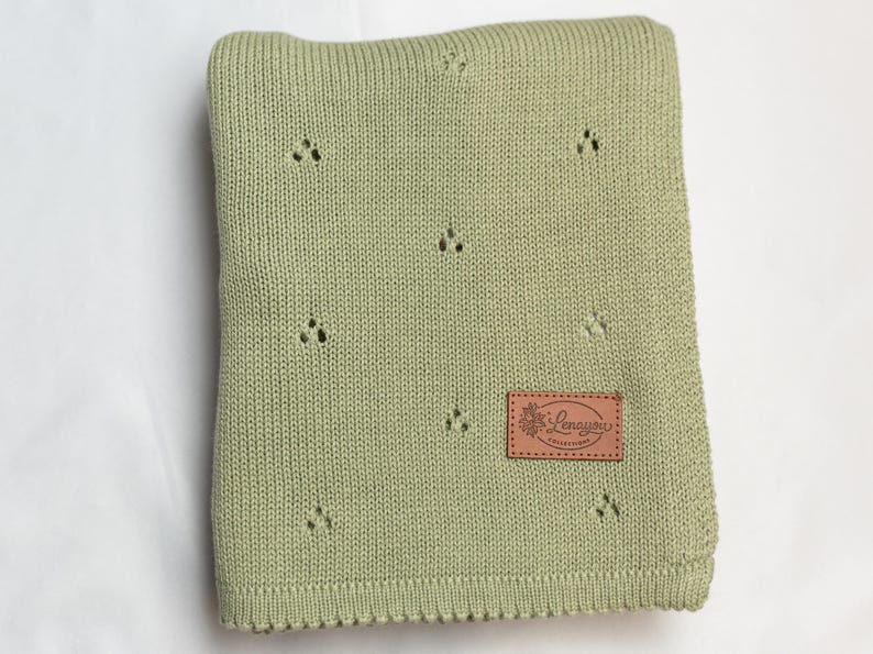 May include: A sage green knitted blanket featuring a repeating pattern of small, three-hole eyelet clusters. A rectangular brown leather-like patch is stitched onto the lower right corner, displaying the embossed text Lenayou Collections.