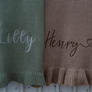 May include: Two knitted baby blankets, one sage green and one tan, with embroidered names. The sage green blanket has the name "Lilly" in white script. The tan blanket has the name "Henry" in brown script with a heart symbol.