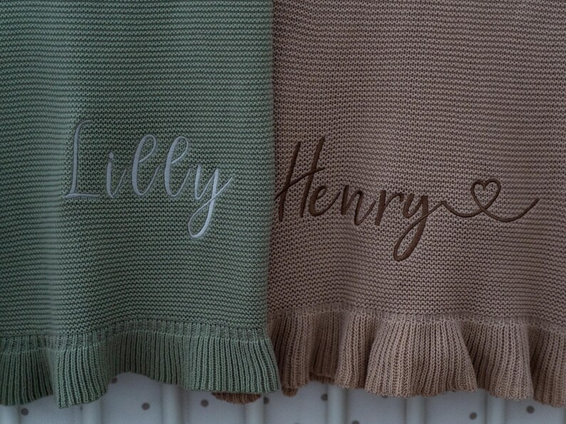 May include: Two knitted baby blankets, one sage green and one tan, with embroidered names. The sage green blanket has the name "Lilly" in white script. The tan blanket has the name "Henry" in brown script with a heart symbol.