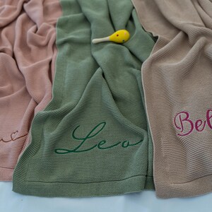 May include: Three knitted baby blankets in shades of pink, green, and tan. Each blanket has a name embroidered in a contrasting color: Alya, Leo, and Bella. A yellow and white maraca rests on the green blanket.