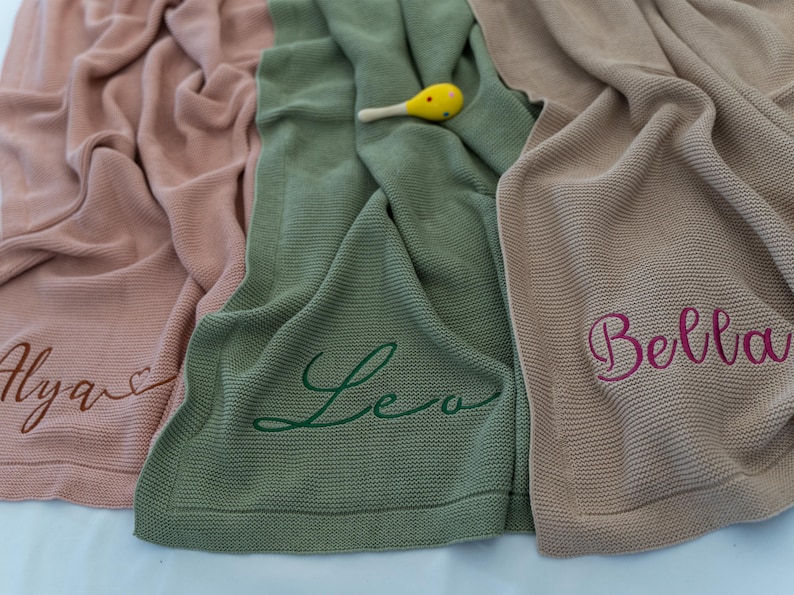 May include: Three knitted baby blankets in shades of pink, green, and tan. Each blanket has a name embroidered in a contrasting color: Alya, Leo, and Bella. A yellow and white maraca rests on the green blanket.