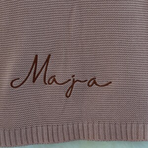 May include: A dusty rose knitted blanket with the name "Maja" embroidered in a darker brown script. The blanket has a ribbed border and a textured appearance.