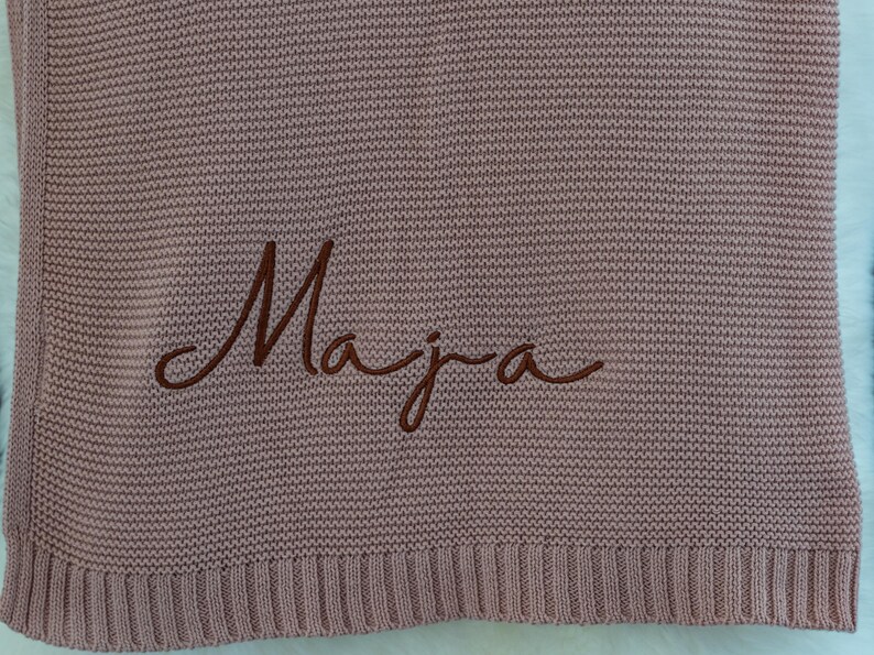 May include: A dusty rose knitted blanket with the name "Maja" embroidered in a darker brown script. The blanket has a ribbed border and a textured appearance.