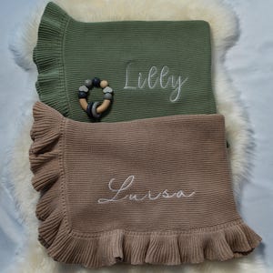 May include: Two baby blankets, one olive green and one tan, with ruffled edges and embroidered names. A wooden and silicone teething ring rests on the green blanket. The blanket's names are "Lilly" and "Luisa".