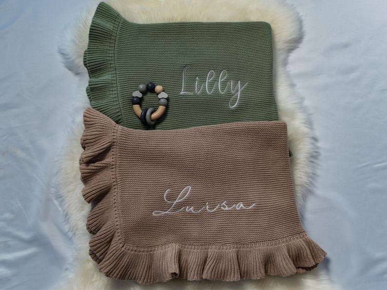 May include: Two baby blankets, one olive green and one tan, with ruffled edges and embroidered names. A wooden and silicone teething ring rests on the green blanket. The blanket's names are "Lilly" and "Luisa".