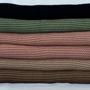 May include: A stack of knitted fabric rolls in various colors. The colors include black, olive green, two shades of pink, and two shades of brown. The fabric has a textured, woven appearance, and the rolls are neatly stacked.