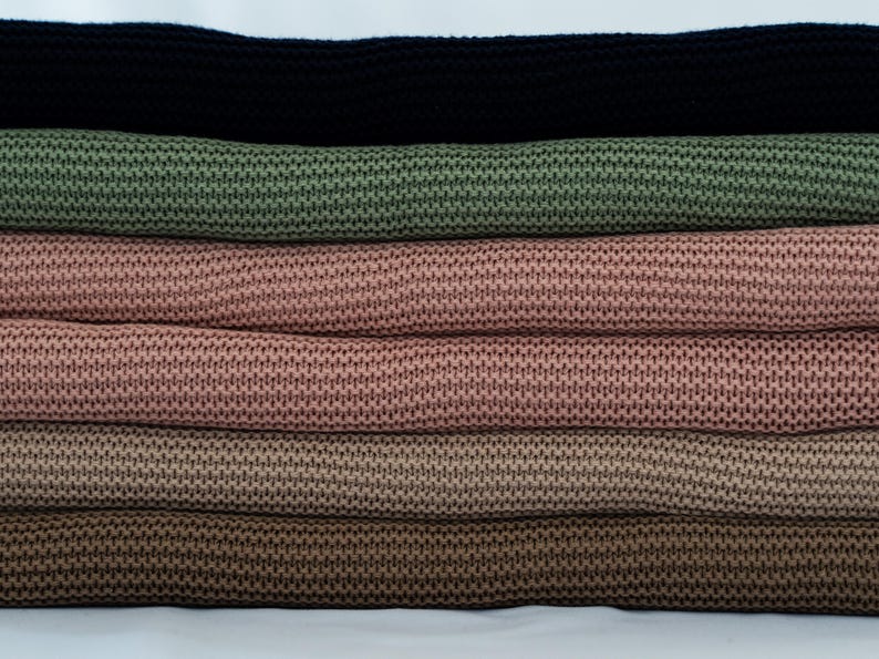 May include: A stack of knitted fabric rolls in various colors. The colors include black, olive green, two shades of pink, and two shades of brown. The fabric has a textured, woven appearance, and the rolls are neatly stacked.