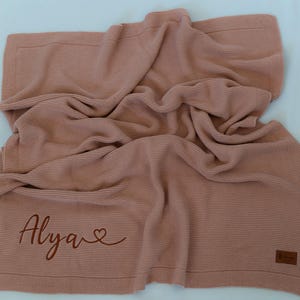 May include: A dusty rose knitted blanket with the name "Alya" and a heart symbol embroidered on the corner. The blanket has a textured appearance and a rectangular shape. A small brown label is on the bottom right corner.