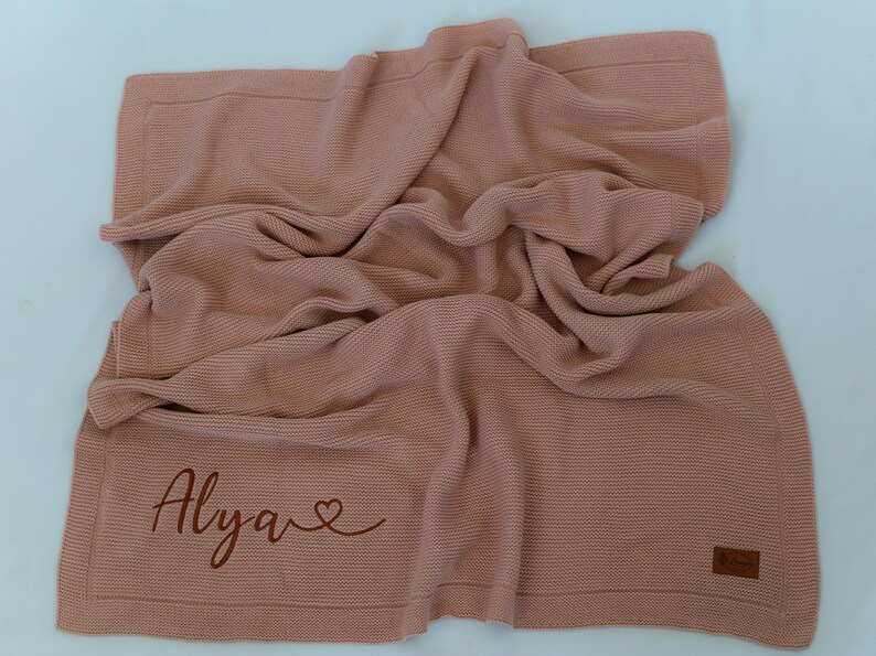 May include: A dusty rose knitted blanket with the name "Alya" and a heart symbol embroidered on the corner. The blanket has a textured appearance and a rectangular shape. A small brown label is on the bottom right corner.