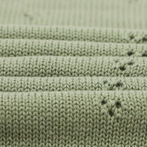 May include: A sage green knit fabric featuring a ribbed texture and a repeating pattern of small, delicate eyelet holes arranged in a subtle, spaced-out design. The material is folded in horizontal layers, showcasing the consistent stitch detail.