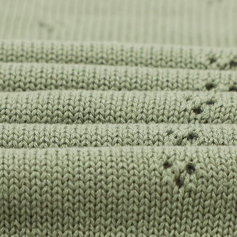 May include: A sage green knit fabric featuring a ribbed texture and a repeating pattern of small, delicate eyelet holes arranged in a subtle, spaced-out design. The material is folded in horizontal layers, showcasing the consistent stitch detail.
