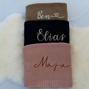 May include: Three personalized knitted blankets in shades of brown, navy, and pink are stacked on a white fluffy surface. Each blanket features a name embroidered in a complementary color: "Ben", "Elias", and "Maja".