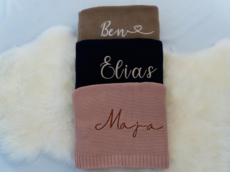 May include: Three personalized knitted blankets in shades of brown, navy, and pink are stacked on a white fluffy surface. Each blanket features a name embroidered in a complementary color: "Ben", "Elias", and "Maja".