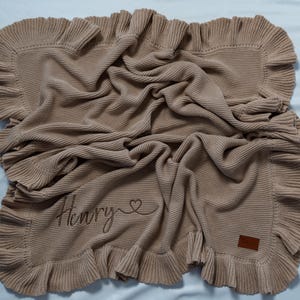 May include: A tan knitted blanket with a ruffled edge. The blanket has the name "Henry" embroidered on it, along with a heart symbol. A small brown leather tag is sewn on the corner.
