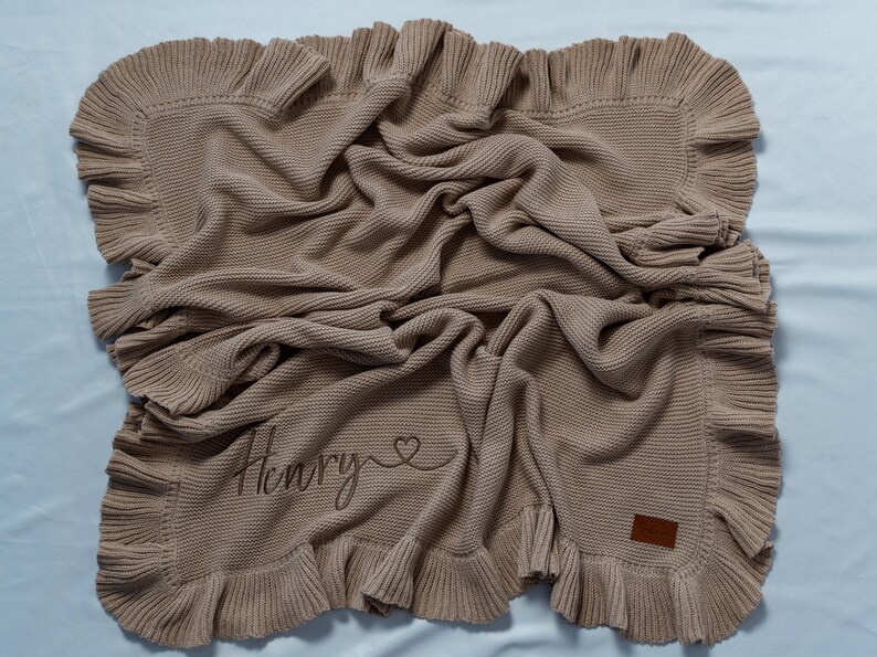 May include: A tan knitted blanket with a ruffled edge. The blanket has the name "Henry" embroidered on it, along with a heart symbol. A small brown leather tag is sewn on the corner.